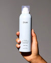 A hand holding a white aerosol can of OUAI Super Dry Shampoo against a solid gray background.