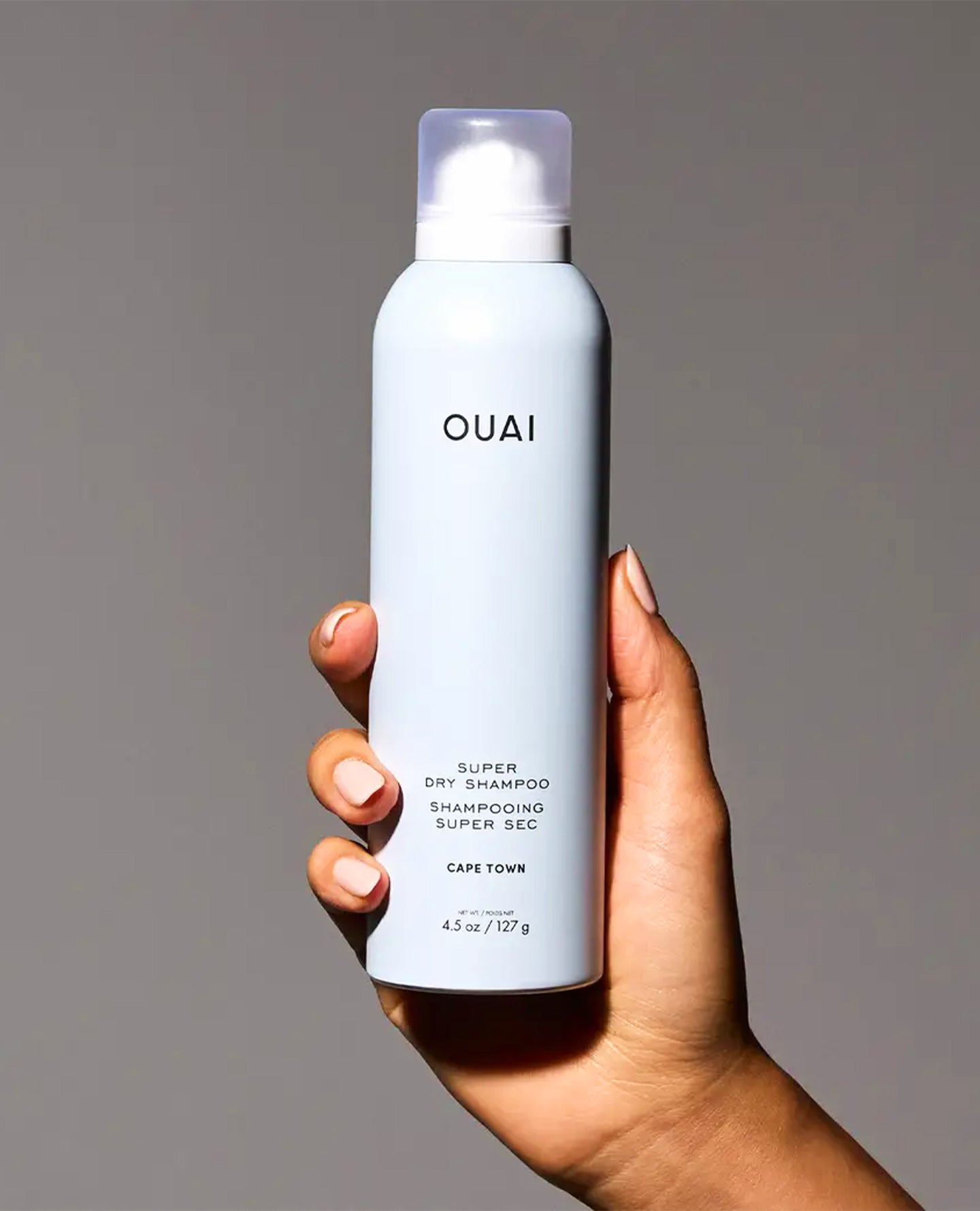 A hand holding a white aerosol can of OUAI Super Dry Shampoo against a solid gray background.