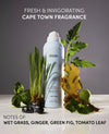 OUAI Fresh & Invigorating Cape Town Fragrance Super Shampoo bottle styled with fresh ingredients including wet grass, ginger, basil, and green fig on a white surface against a dark gray background.