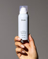 A hand with manicured nails holds a white bottle of OUAI Super Dry Shampoo against a solid gray background.