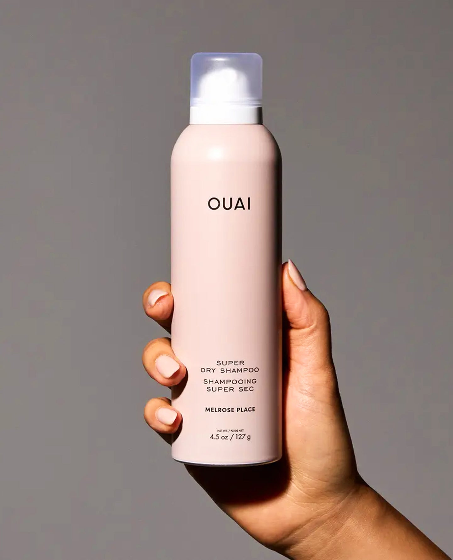A hand holding a matte blush pink bottle of OUAI Super Dry Shampoo against a solid gray background.