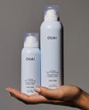 Two OUAI haircare bottles held in a person's hand against a plain gray studio background. The smaller bottle is pale icy blue and the larger bottle is white, both with black OUAI branding and black text.