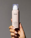 A hand with manicured nails holds a bottle of OUAI Super Dry Shampoo in the Melrose Place scent against a solid gray background.