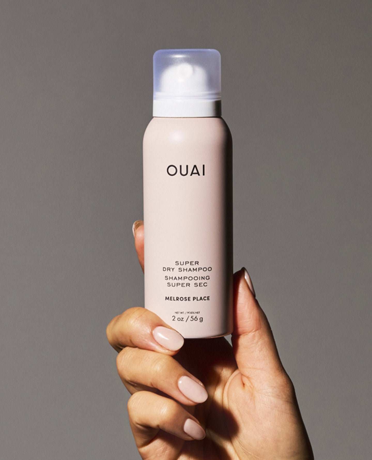A hand with manicured nails holds a bottle of OUAI Super Dry Shampoo in the Melrose Place scent against a solid gray background.