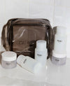 A set of OUAI clear empty travel bottles including a clear zippered cosmetic bag and several frosted bottles and jars with white caps, arranged on a glossy white surface against a white tiled background.