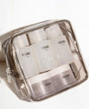 A clear zippered cosmetic pouch containing four OUAI empty travel bottles and two jars, photographed on a white tiled surface.