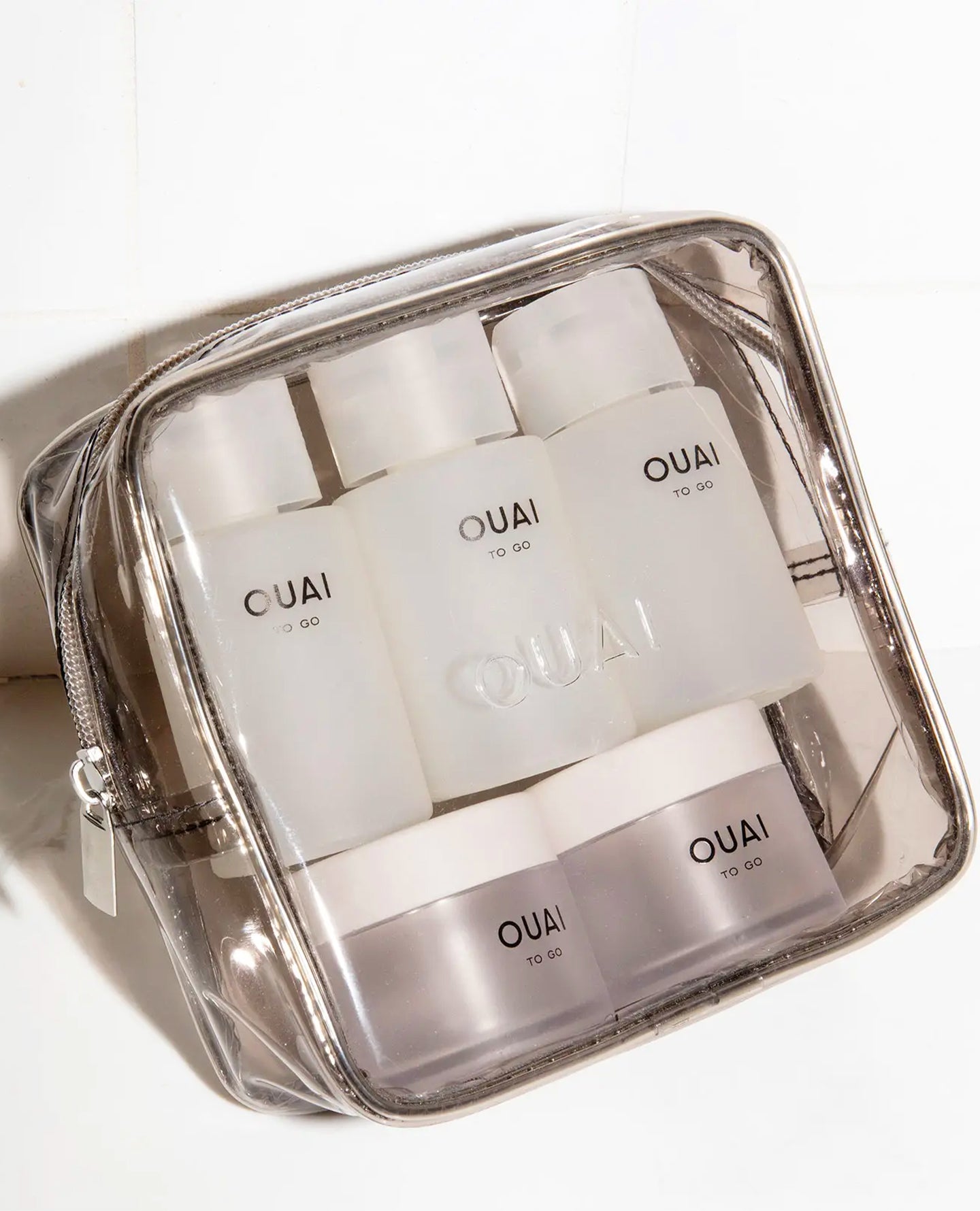 A clear zippered cosmetic pouch containing four OUAI empty travel bottles and two jars, photographed on a white tiled surface.