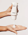 A hand holds a white OUAI Tuck-Max Hair Grows Fast cream-colored squeeze bottle, dispensing thick white hair cream into another hand against a light gray background.