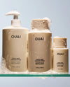 Three OUAI Thick Hair Conditioner bottles in beige and cream tones, shown with white foam on a glass shelf against a pale blue background.