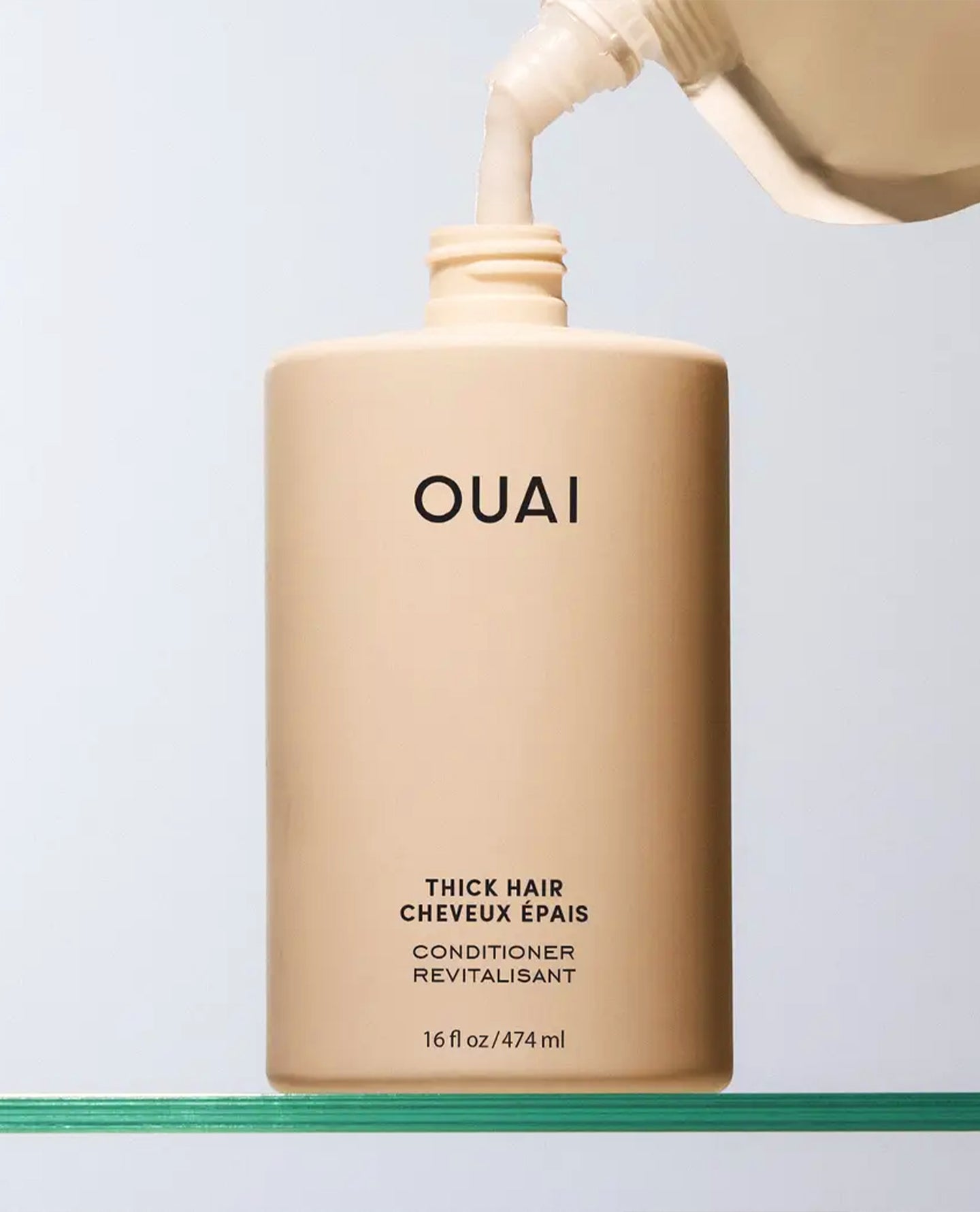 OUAI Thick Hair Conditioner bottle in a warm beige shade, with conditioner being poured from a matching dispenser above it against a light gray background.
