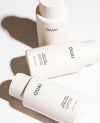 Three OUAI skincare bottles arranged diagonally on a white background with soft shadows. The bottles are a warm off-white/cream color with black text, showing the OUAI brand name and product names for thickening facial gel and hydrating face gel.
