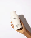 A hand holding a white bottle of OUAI Thick hair conditioner against a clean white background with a soft shadow.