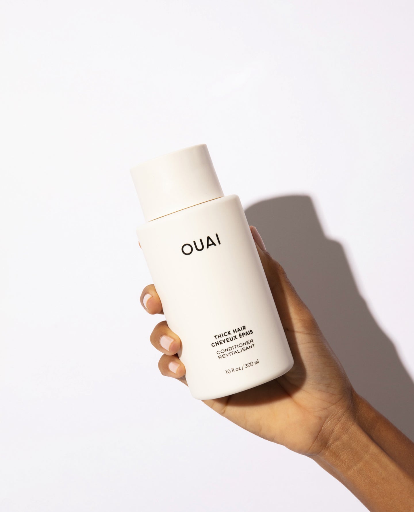 A hand holding a white bottle of OUAI Thick hair conditioner against a clean white background with a soft shadow.