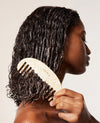 A woman with wet, dark curly hair holds a cream-colored carved wooden comb against her hair. 