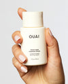 A hand with manicured nails holds a small white bottle of OUAI Thick Hair Conditioner against a light gray background.