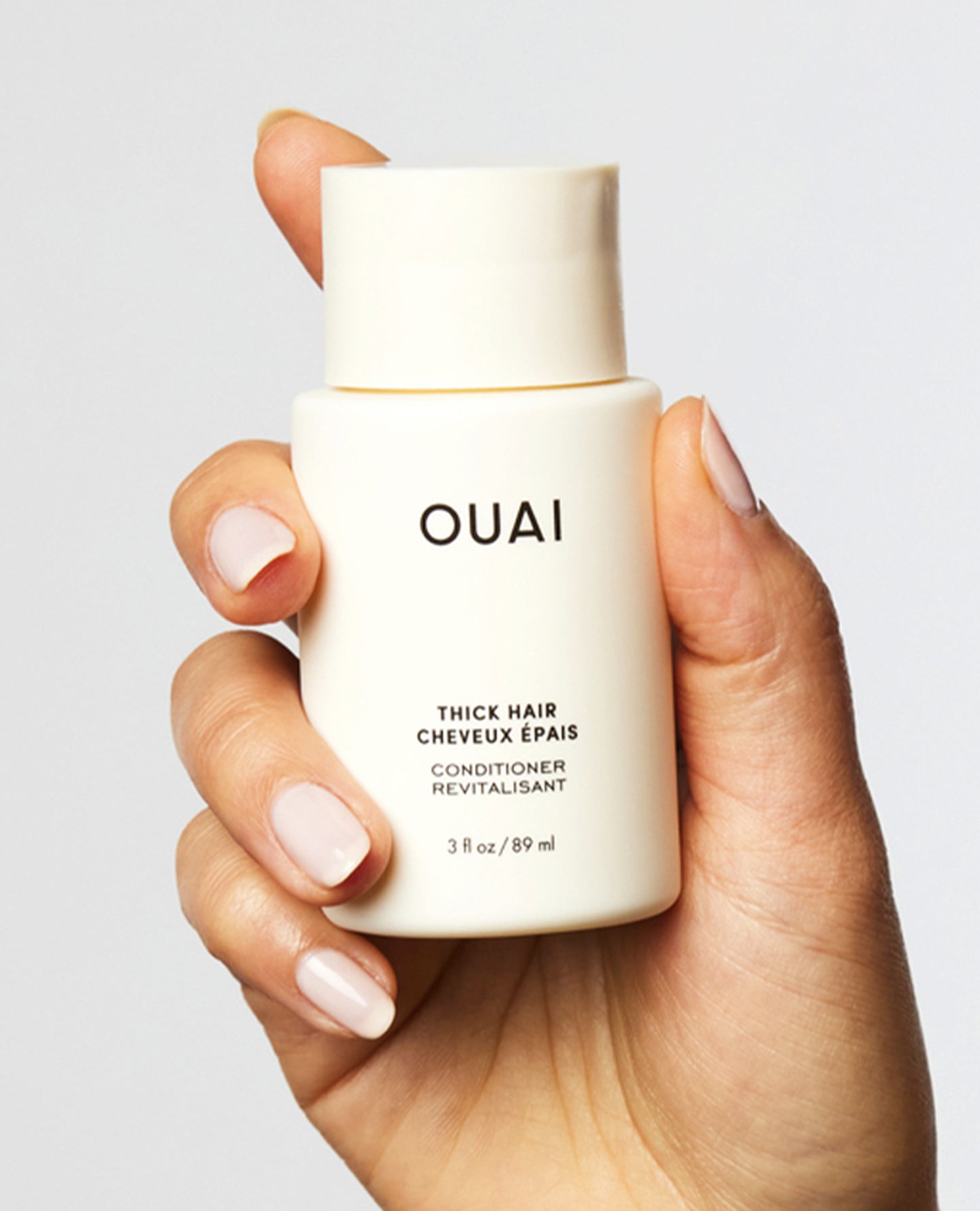 A hand with manicured nails holds a small white bottle of OUAI Thick Hair Conditioner against a light gray background.