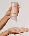A hand holds a frosted bottle of OUAI Ceramide Epa Hand Cream, with the cream being squeezed into the other hand against a plain light background.