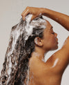 A close-up lifestyle shot of a woman with long, dark, curly hair washing it with foamy shampoo, set against a plain light gray background.