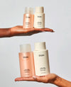 Two hands holding OUAI hair care bottles against a light gray studio background. The top hand holds a peach-toned face air conditioner and a cream-colored conditioner bottle.