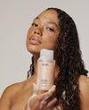 A woman with wet curly hair and freckles holds a bottle of OUAI No. 08 Hydrating Cleanser against a plain light gray background.