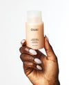 A hand with long, manicured light pink nails holds a bottle of OUAI Thick Hair Shampoo against a clean white background.