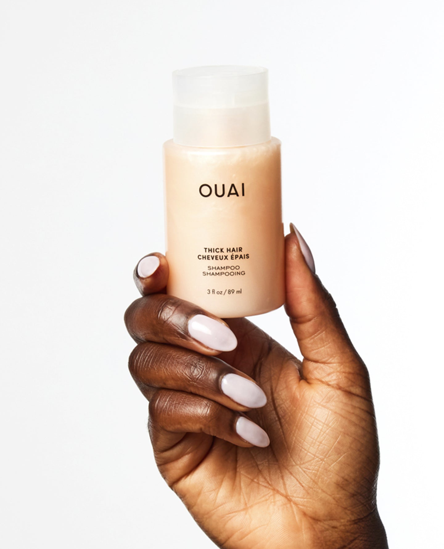 A hand with long, manicured light pink nails holds a bottle of OUAI Thick Hair Shampoo against a clean white background.