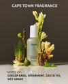 A bottle of OUAI thickening spray centered on a reflective surface with botanical props including green grass, flowers, fig, and ginger. Text at the top reads 'CAPE TOWN FRAGRANCE' and text at the bottom lists notes: ginger basil, spearmint, green fig, wet grass.