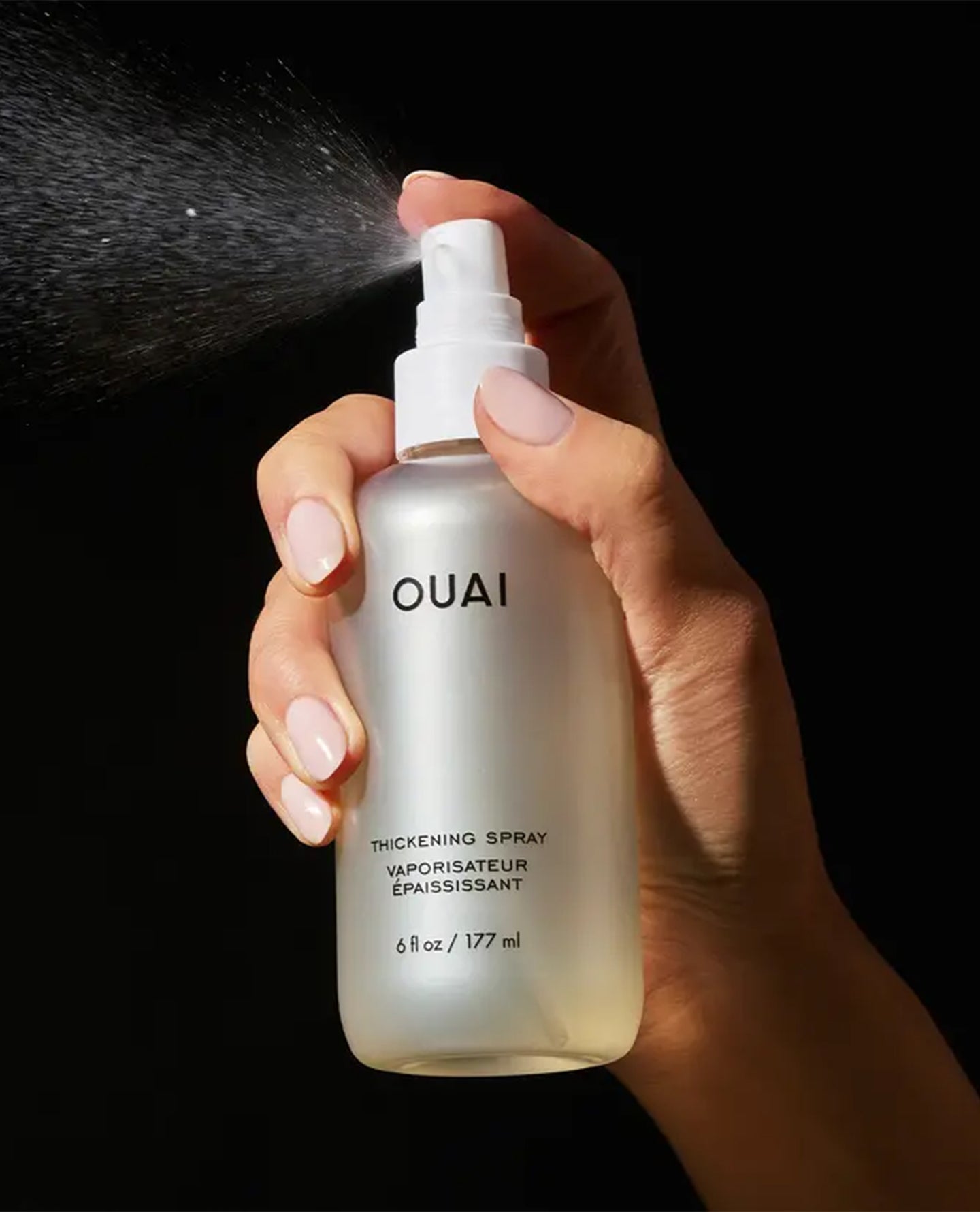 A hand holding a frosted glass spray bottle labeled OUAI Thickening Spray, with the spray misting out against a solid black background.