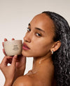 A woman with wet, styled dark hair holds a beige jar of OUAI Shea Hair Repair Mask next to her face, with a white cream visible on her shoulder against a warm beige background.