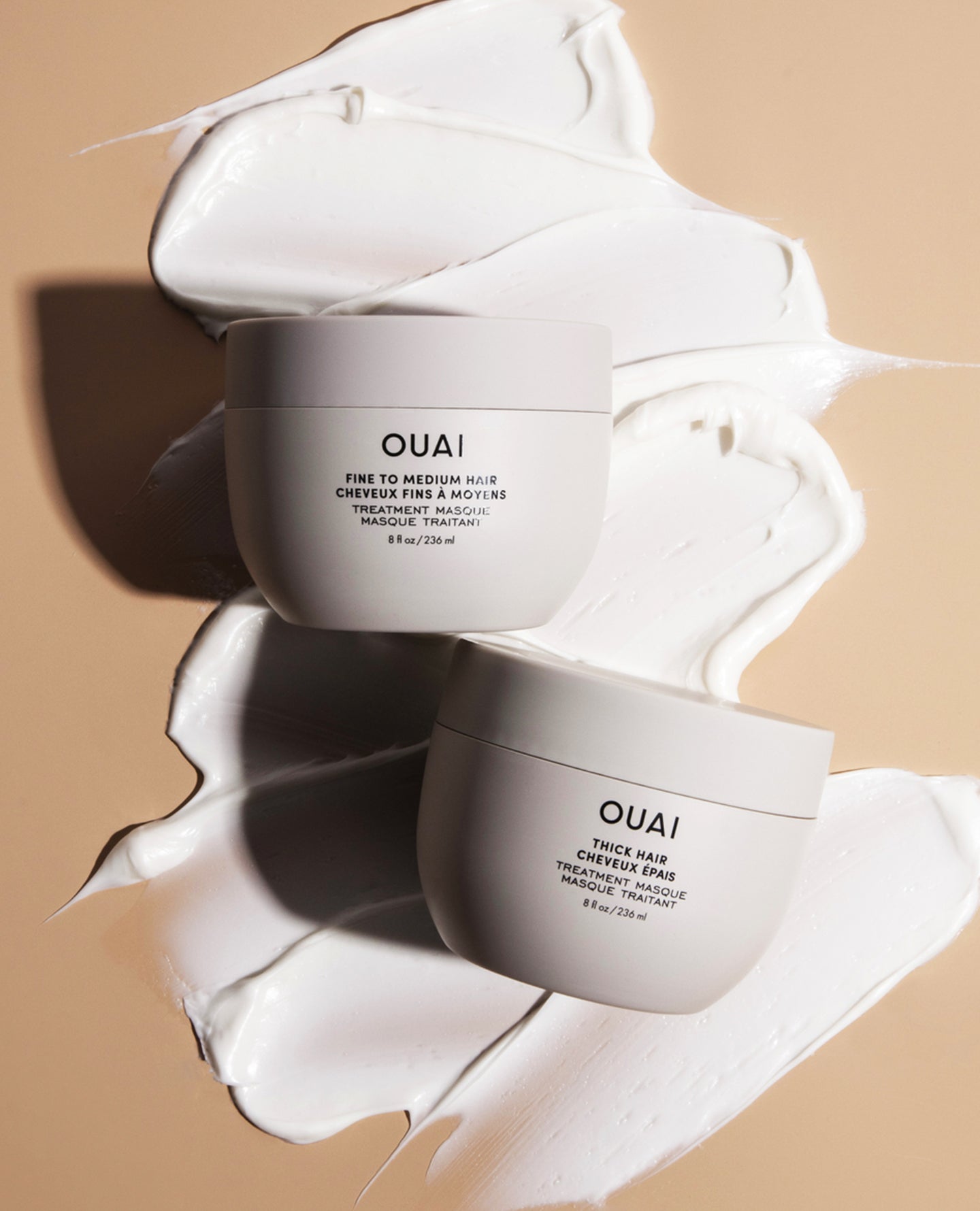 Two OUAI skincare cream jars arranged on a beige background with white cream swatches behind them. The top jar reads 'OUAI Fine to Medium AHA Cheveux Fine & Modifying Treatment Cream' and the bottom jar reads 'OUAI Thick AHA Exfoliating Treatment Cream.