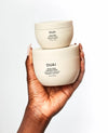 A hand holds two cream-colored OUAI cosmetic jars stacked vertically against a clean white background. The larger jar in the foreground reads 'OUAI THICK HAIR CHEESE SHEEP THICKENER TREATMENT SHAPING & THICKENING AGENT', and the smaller jar behind it reads 'OUAI THICK HAIR CHEESE SHEEP'.