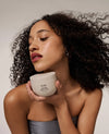 A woman with voluminous dark curly hair holds a white jar of OUAI Hair & Skin Cream against her chest, wearing a gray strapless top against a neutral light gray background.