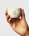 A hand holding a cream-colored jar of OUAI Thick Hair Treatment against a light gray background.