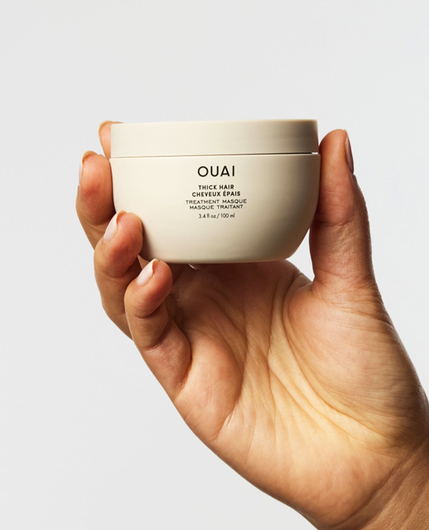A hand holding a cream-colored jar of OUAI Thick Hair Treatment against a light gray background.