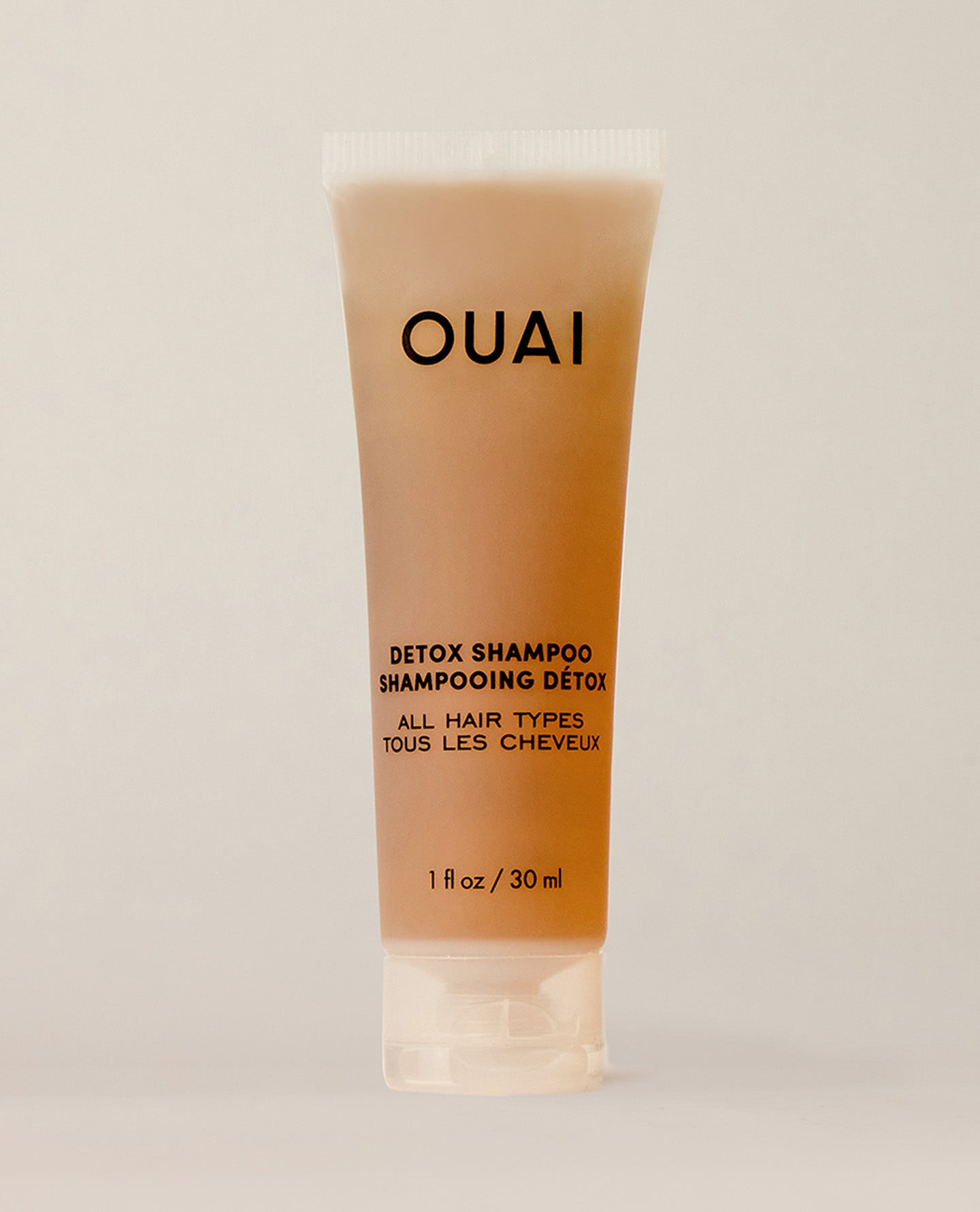 A travel-sized amber shampoo tube stands upright on a softly lit cream surface. Text: “OUAI”, “DETOX SHAMPOO”, “SHAMPOOING DÉTOX”, “ALL HAIR TYPES”, “TOUS LES CHEVEUX”, “1 fl oz / 30 ml”.
