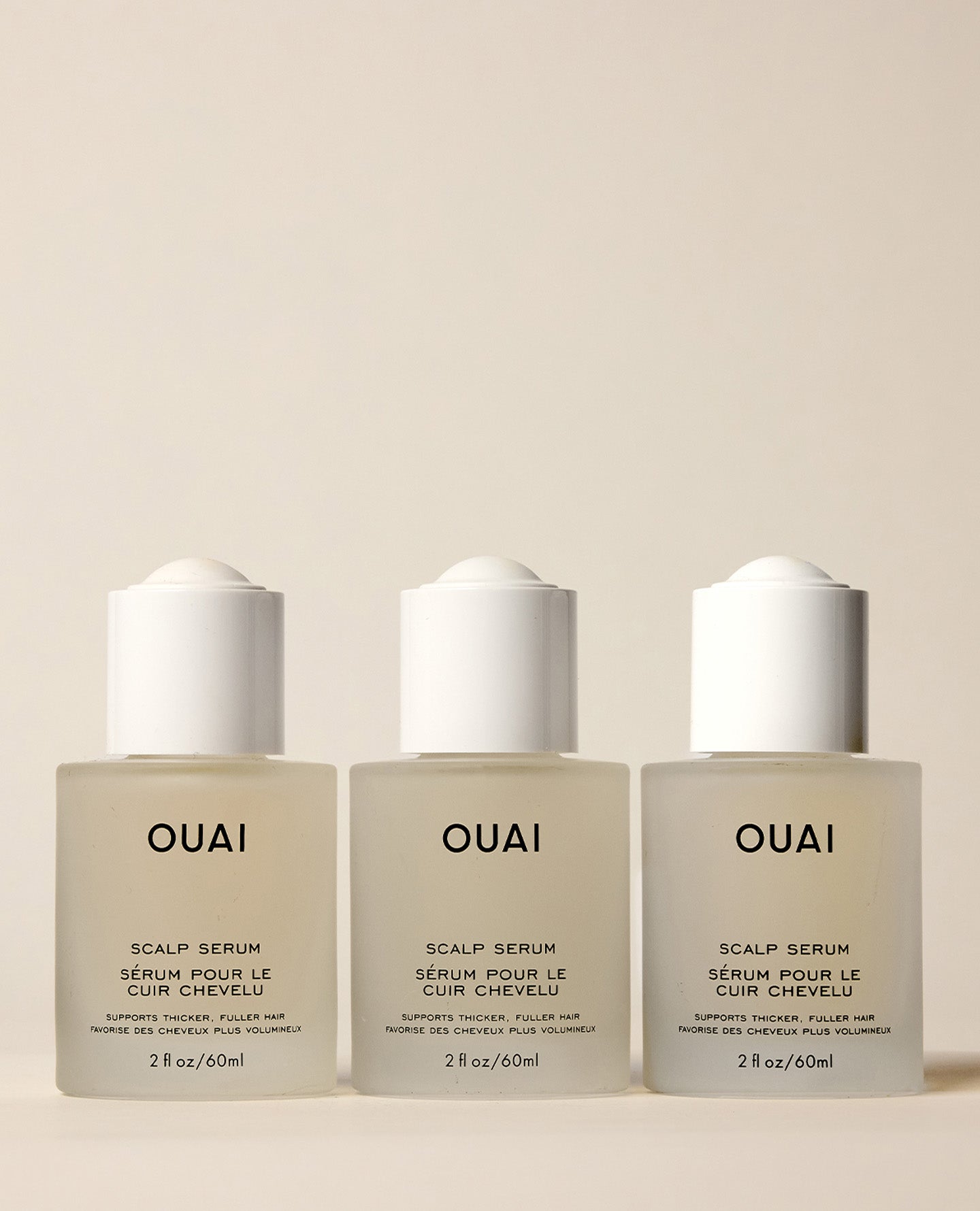 Three identical frosted OUAI Scalp Serum bottles stand side by side on a cream surface, presenting a 90-day supply beneath white applicator caps.