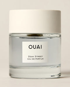 A short clear perfume bottle containing pale blue liquid stands on a cream studio surface beneath a wide white cap. Text: “OUAI”, “DEAN STREET”, “EAU DE PARFUM”.