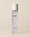 A slim clear OUAI Laurel Canyon eau de parfum bottle containing pale pink liquid stands upright against a warm, softly lit cream studio background.