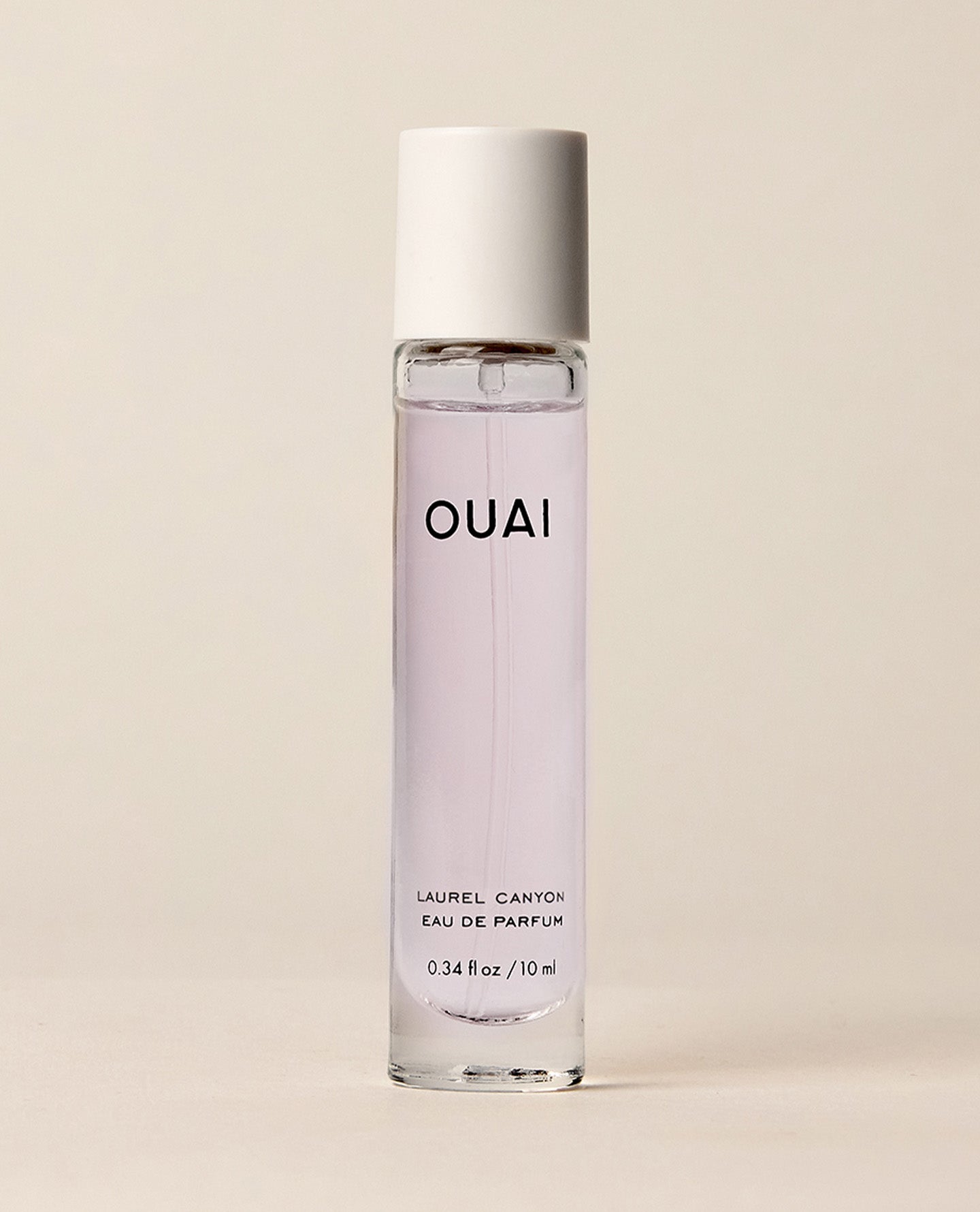 A slim clear OUAI Laurel Canyon eau de parfum bottle containing pale pink liquid stands upright against a warm, softly lit cream studio background.