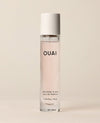 A slim travel perfume bottle containing pale pink liquid stands upright against a softly lit cream backdrop. Text: “OUAI”, “MELROSE PLACE”, “EAU DE PARFUM”, “0.34 fl oz / 10 ml”.