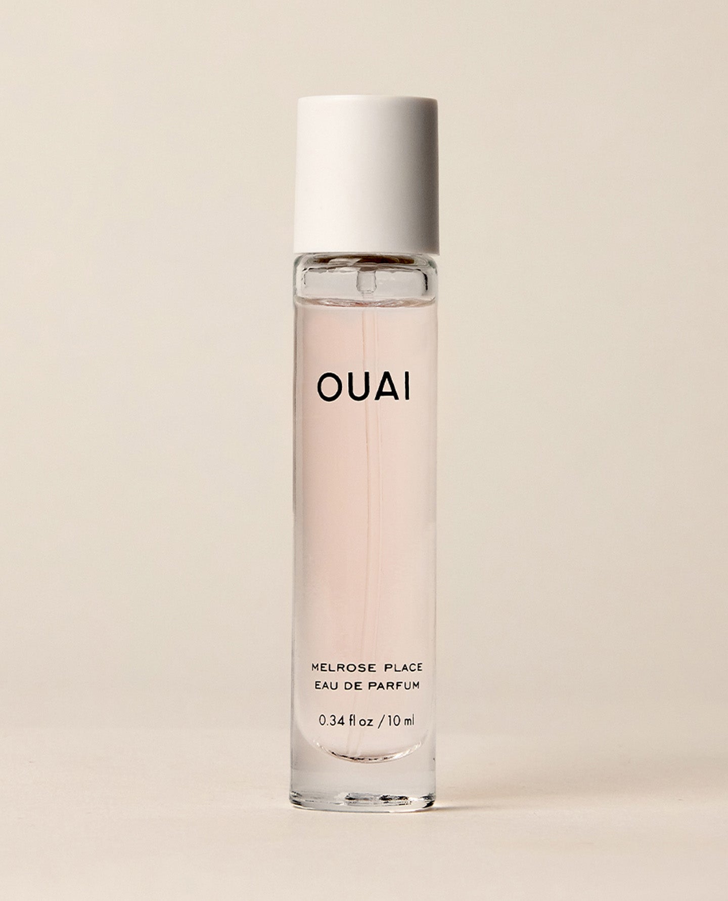 A slim travel perfume bottle containing pale pink liquid stands upright against a softly lit cream backdrop. Text: “OUAI”, “MELROSE PLACE”, “EAU DE PARFUM”, “0.34 fl oz / 10 ml”.
