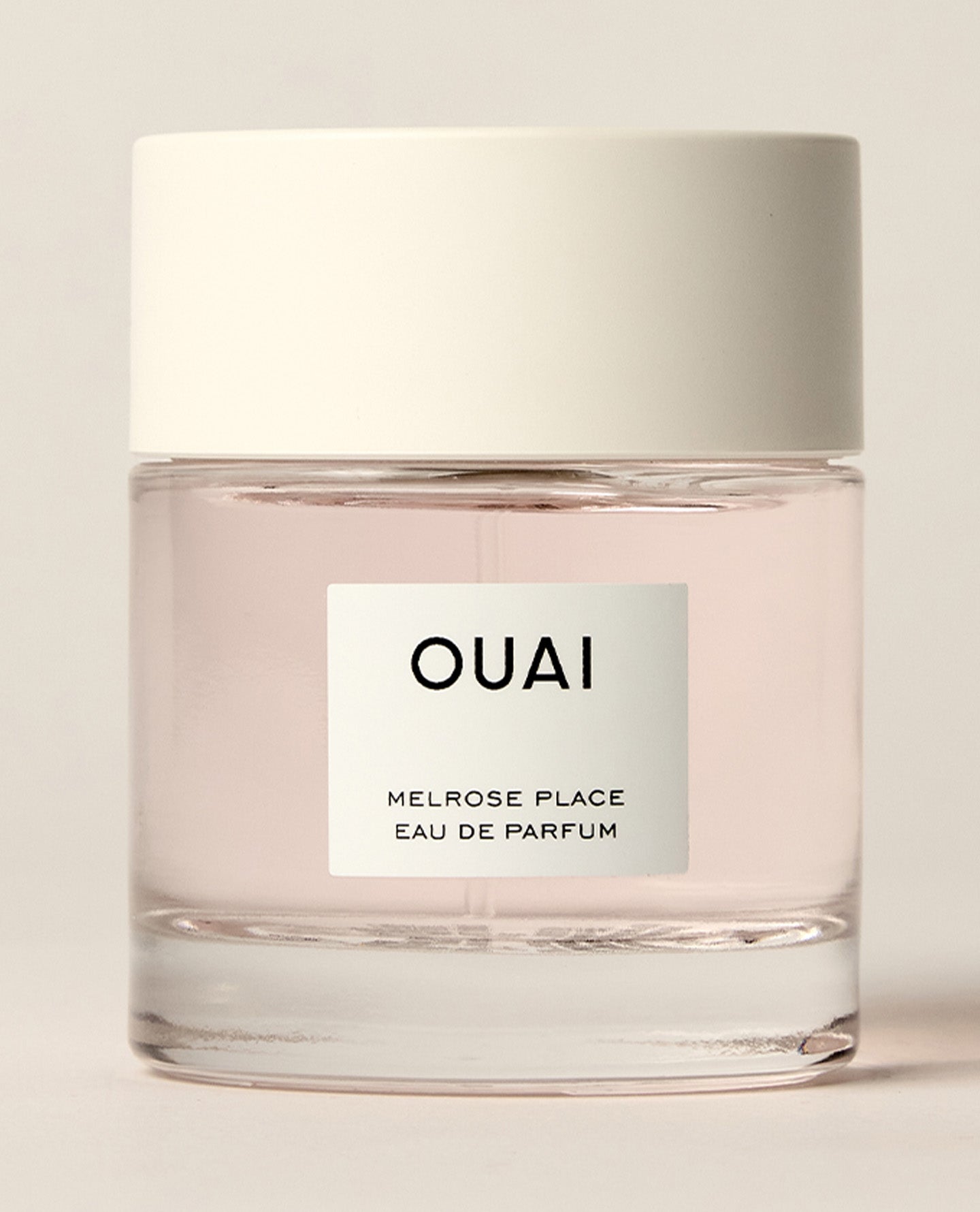 A short clear perfume bottle containing pale pink liquid stands on a warm cream studio surface beneath a wide white cap. Text: “OUAI”, “MELROSE PLACE”, “EAU DE PARFUM”.