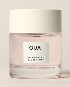 A short clear perfume bottle containing pale pink liquid stands on a warm cream studio surface beneath a wide white cap. Text: “OUAI”, “MELROSE PLACE”, “EAU DE PARFUM”.
