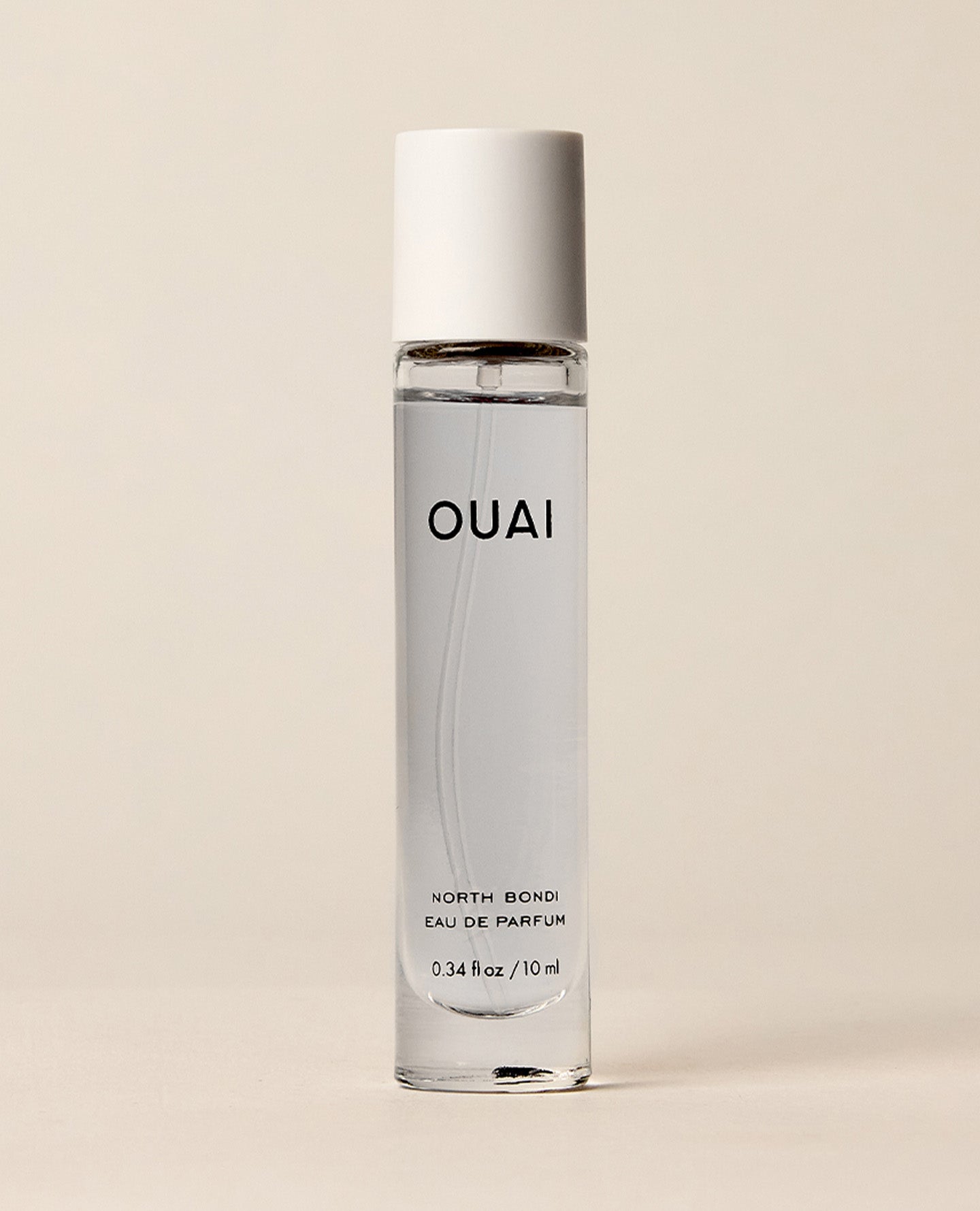 A slim travel perfume bottle containing pale blue liquid stands upright against a softly lit cream backdrop. Text: “OUAI”, “NORTH BONDI”, “EAU DE PARFUM”, “0.34 fl oz / 10 ml”.