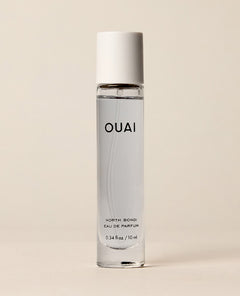 A slim travel perfume bottle containing pale blue liquid stands upright against a softly lit cream backdrop. Text: “OUAI”, “NORTH BONDI”, “EAU DE PARFUM”, “0.34 fl oz / 10 ml”.
