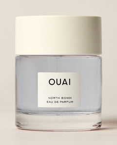 A short clear perfume bottle containing pale blue liquid stands on a cream studio surface beneath a wide white cap. Text: “OUAI”, “DEAN STREET”, “EAU DE PARFUM”.