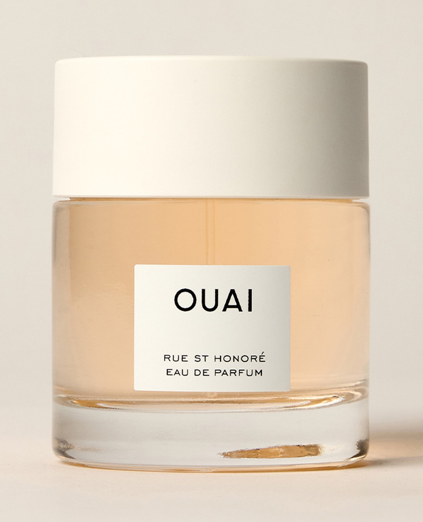 A short clear perfume bottle containing pale peach liquid stands beneath a wide white cap against a cream background. Text: “OUAI”, “RUE ST HONORÉ”, “EAU DE PARFUM”.