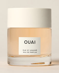 A short clear perfume bottle containing pale peach liquid stands beneath a wide white cap against a cream background. Text: “OUAI”, “RUE ST HONORÉ”, “EAU DE PARFUM”.