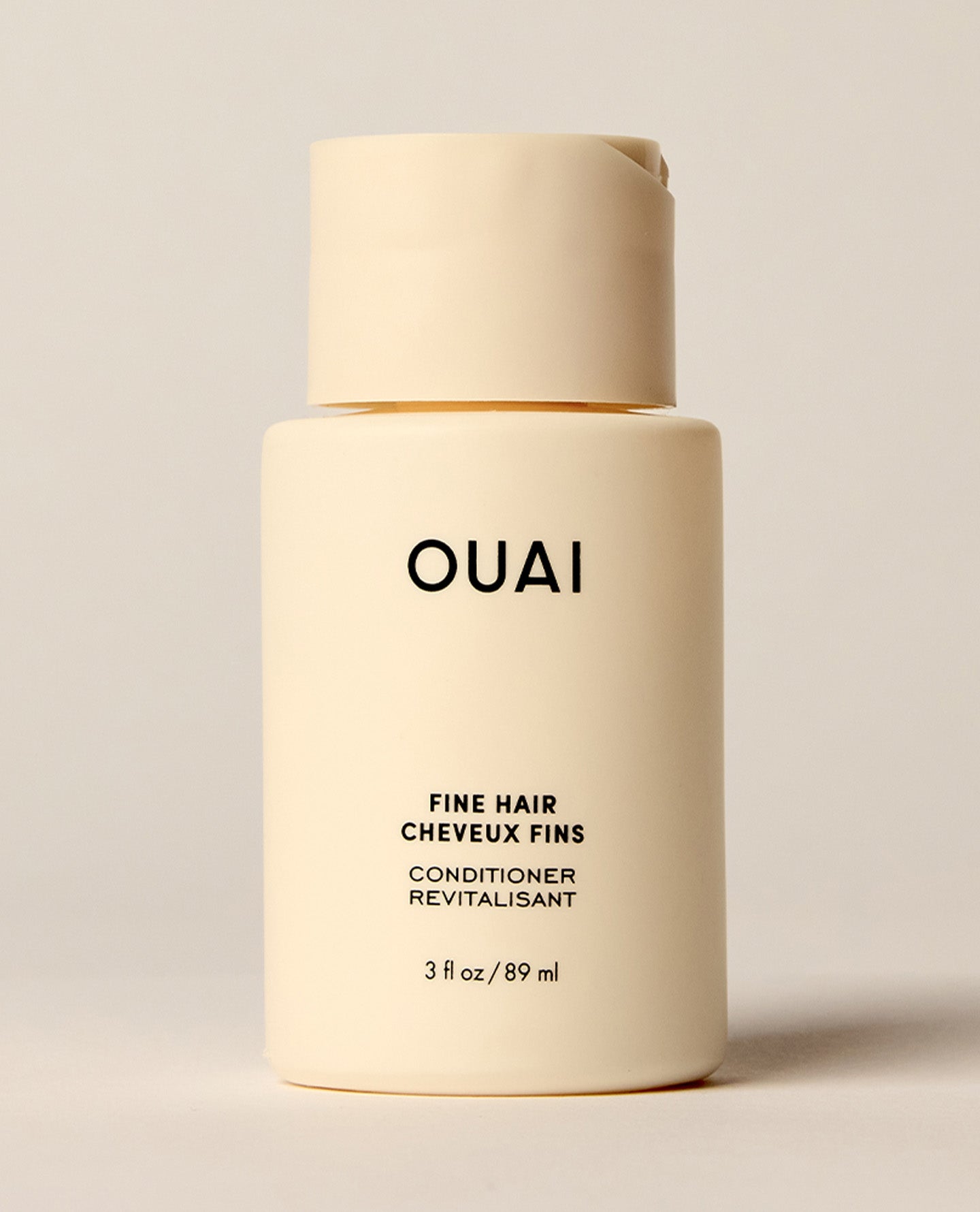 A travel-sized ivory OUAI Fine Hair Conditioner bottle stands upright against a softly lit cream background. Text: “OUAI”, “FINE HAIR”, “CHEVEUX FINS”, “CONDITIONER”, “REVITALISANT”, “3 fl oz / 89 ml”.