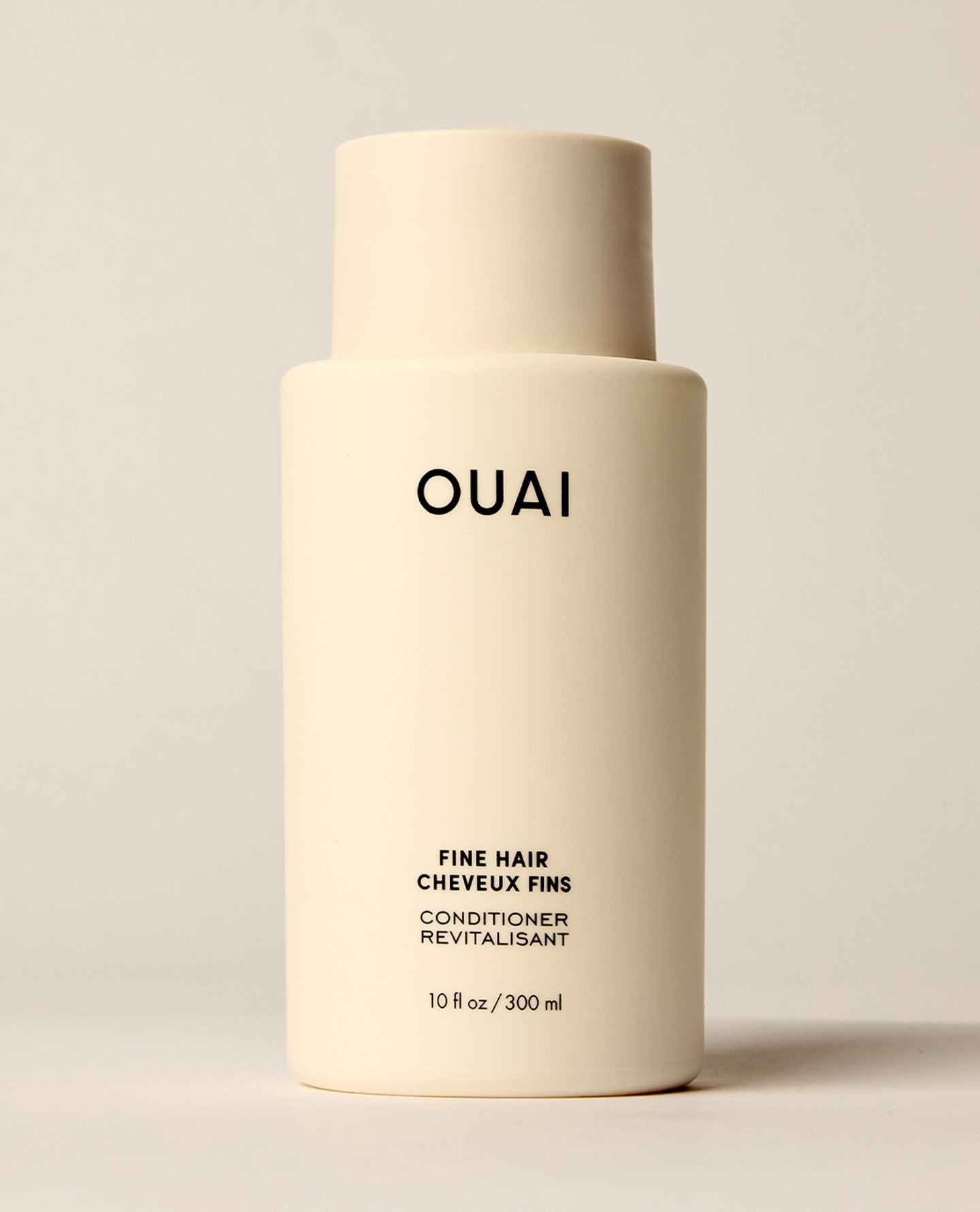 An ivory OUAI Fine Hair Conditioner bottle stands upright on a softly lit cream studio surface. Text: “OUAI”, “FINE HAIR”, “CHEVEUX FINS”, “CONDITIONER”, “REVITALISANT”, “10 fl oz / 300 ml”.